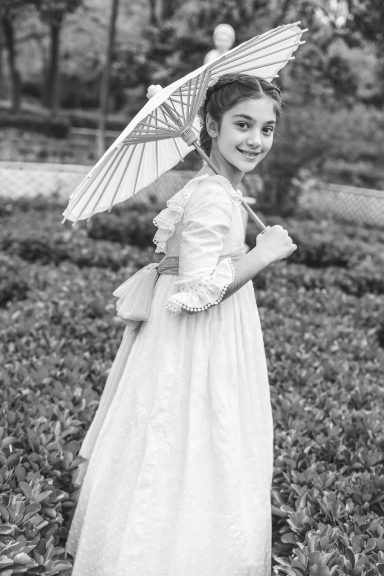 Niña sonriente en un jardín, vestida con un vestido blanco y sosteniendo un paraguas.
